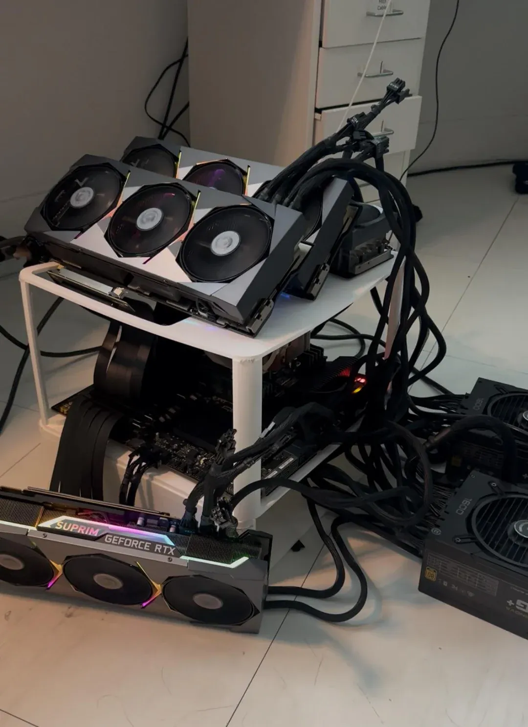 Open-air AI rig built around a small white IKEA table, with MSI Suprim RTX 3090 cards on top and on the floor, the motherboard underneath, and several power supplies with thick cable bundles beside it.
